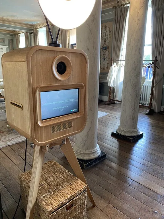 A premium photo booth hire stands on a tripod in an elegant room with marble columns, large windows, wooden floors, and ornate decor. A soft light tops the booth, with a wicker basket beneath—perfect for a sophisticated corporate event experiential.