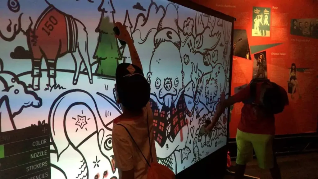 Two children interact with a large, illuminated graffiti wall hire display, colouring whimsical black-and-white drawings with stylus pens in a dimly lit, colourful exhibit space—perfect for a unique corporate event experiential experience.